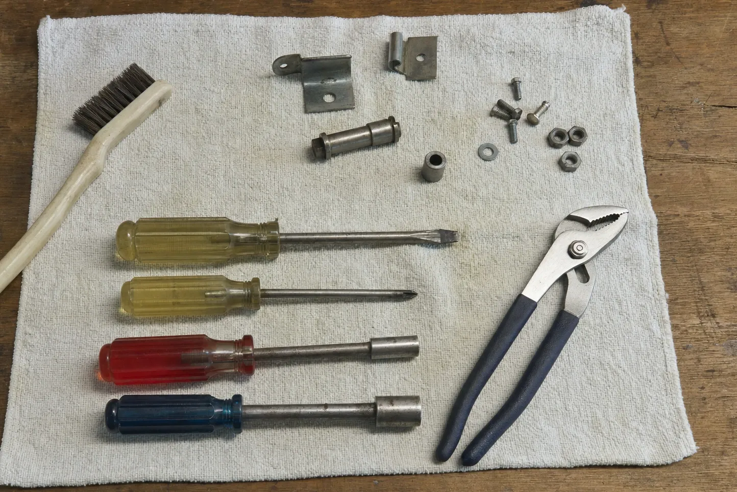 Wire brush, hinge brackets, a roller pin and bushing, nuts and washers on a cloth beside four drivers and slip-joint pliers