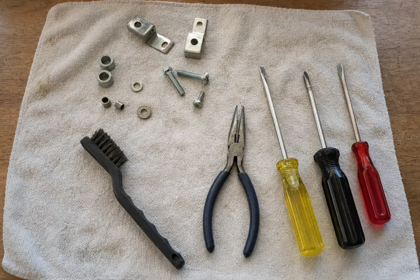 Hinge brackets, bushings, bolts and washers laid out on a cloth beside long-nose pliers and three screwdrivers