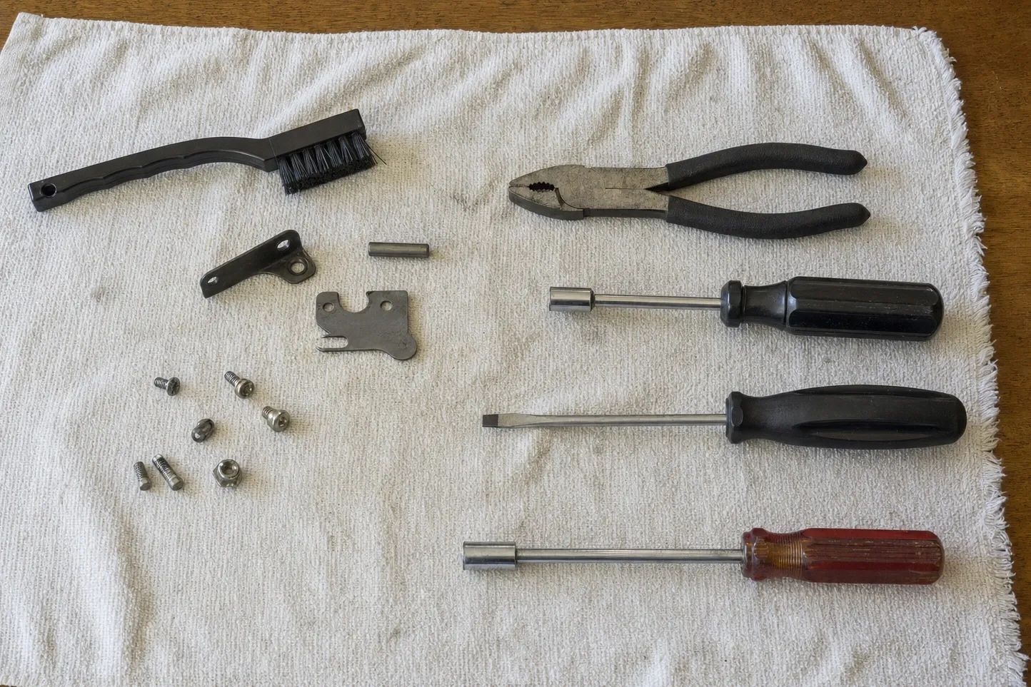A hooked steel latch plate, an angle bracket and a roller pin on a white towel beside a wire brush, slip-joint pliers and three drivers